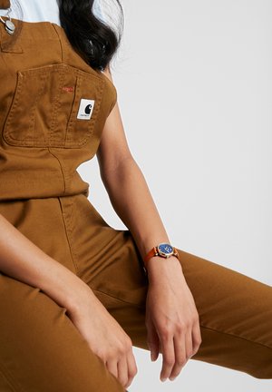Person wearing brown Carhartt overalls and an orange-strapped watch with a blue face, against a plain light gray background.