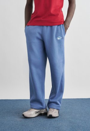 Asmuo, vilkintis šviesiai mėlynus Nike sportinius džinsus, baltus Nike sportbačius ir raudoną marškinėlį, stovintis ant mėlyno kilimo su rankomis kišenėse.