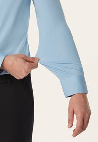 Lichtblauwe overhemd met lange mouwen en een soepele textuur, met een strakke manchette, die aan de mouw wordt getrokken door een hand tegen een neutrale achtergrond.