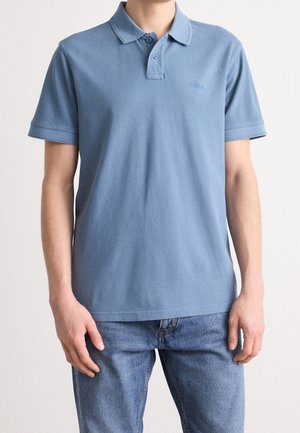 Person iført en lyseblå kortærmet polo og blå jeans, stående med hænderne afslappet ned langs siderne mod en ensfarvet baggrund.