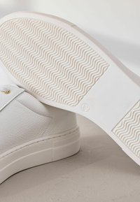 Baskets en cuir blanc avec côté texturé, semelle en caoutchouc plate avec un motif ondulé, et œillets dorés pour les lacets.