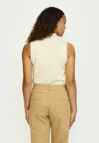 Bela brezrokavna bluza z visokim izrezom, v kombinaciji z bež hlačami, ki imata gladko teksturo in minimalističen dizajn.