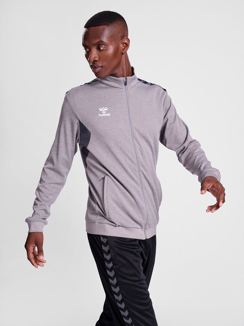 Modèle masculin portant une veste grise à zip et un pantalon noir à motifs, posant avec les bras tendus sur un fond clair uni.