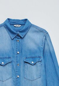 Light blue denim shirt with a collar, front buttons, and two chest pockets. Features silver snap buttons and subtle stitching details.
