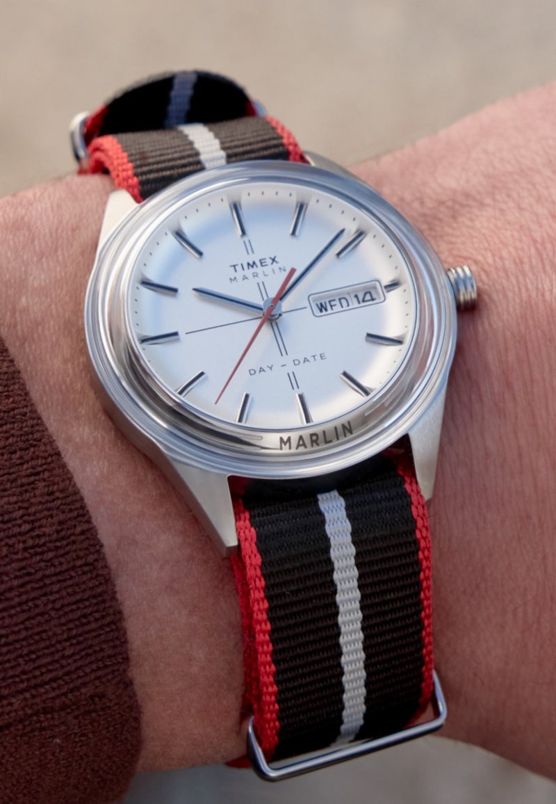 Timex Marlin-horloge met zilverkleurige wijzerplaat, zilveren uurmarkeringen, rode secondewijzer en zwart/rood gestreepte nylon band, voorzien van dag- en datumaanduiding.