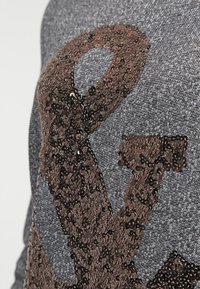 Tessuto grigio con grandi abbellimenti in paillettes marroni a forma di lettere. La texture è liscia e riflettente grazie alle paillettes.