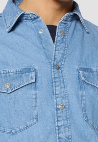 Camicia di jeans azzurro chiaro con design a bottoni, colletto abbattuto, due tasche sul petto e bottoni a snap in tonalità argento. Tessuto con texture visibile.