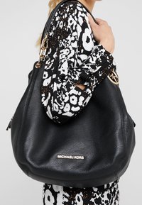 Sort læder hobo-taske med en slap form, med en logo og hardware i guldtoner. Tasken har en tekstureret overflade uden synlige mønstre.