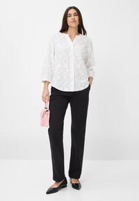 Weiße, floral bestickte Bluse mit Knopfleiste, kombiniert mit schwarzen geraden Hosen und schwarzen spitzen Ballettschuhen. Hält eine kleine rosa Tasche.