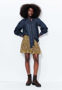 Blouse en denim avec un détail de cou noué, associée à une jupe jaune à motifs. Bottines Chelsea en daim marron. Le mannequin se tient devant un fond blanc.