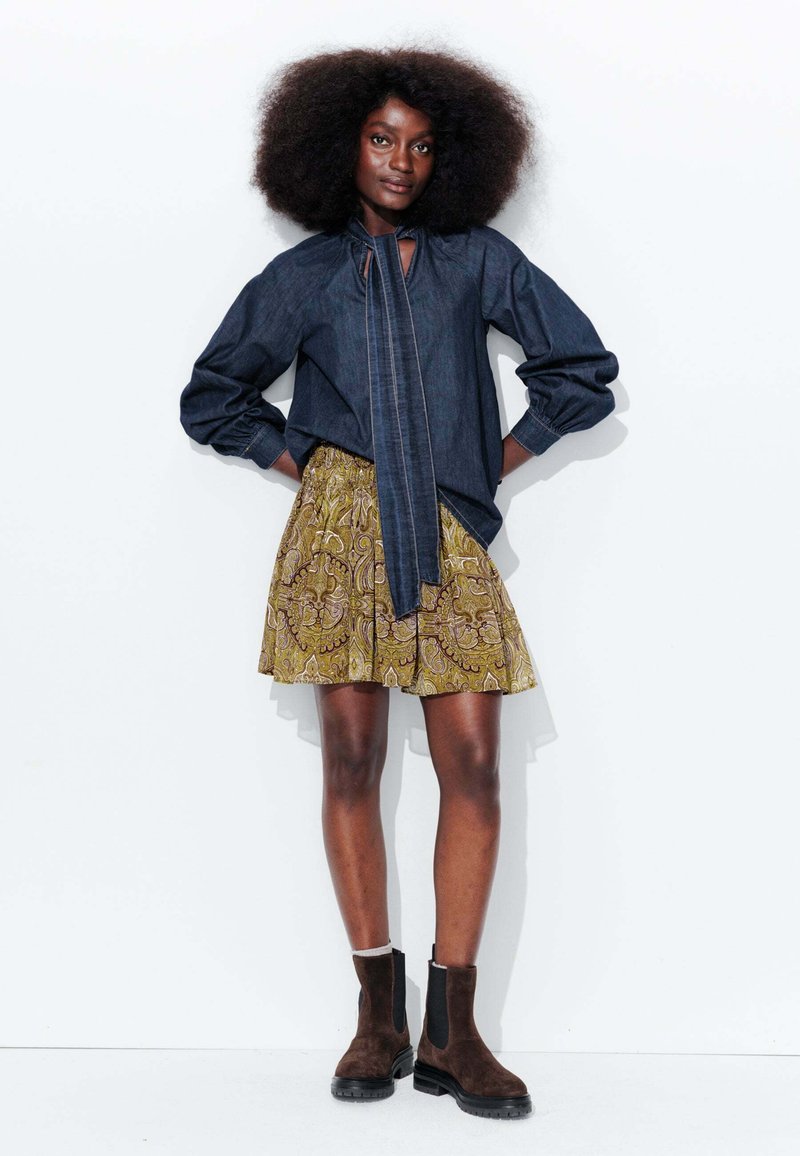 Blouse en denim avec un détail de cou noué, associée à une jupe jaune à motifs. Bottines Chelsea en daim marron. Le mannequin se tient devant un fond blanc.