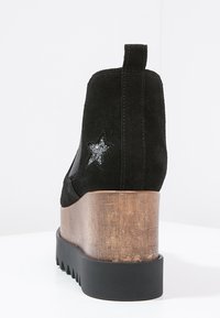 Botín de ante negro con un tacón de cuña de madera marrón, que presenta un detalle de estrella brillante plateada en el lateral y una suela de goma texturizada.