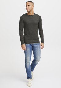 Dark green long-sleeve shirt with a crew neckline, paired with light blue slim-fit jeans and white sneakers, standing against a neutral background.