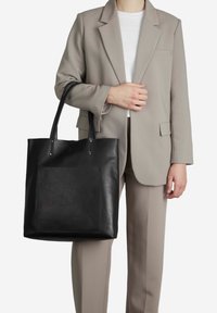 Juodas odinis rankinis krepšys su dviem ilgais rankenomis, lygiu paviršiumi ir priekinėmis išorinėmis kišenėmis. Nešiojamas su šviesiai pilku kostiumu.