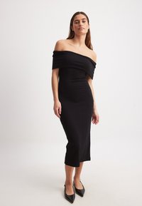 Melnā ribota midi kleita ar atvērtām plecu daļām, pieguļošs siluets un izgriezums apakšmalā, pārī ar melnām norakstu kurpēm ar smailu purngalu.