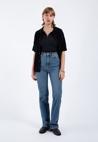 Svart kortärmad skjorta med knappar över en ribbad svart topp, i kombination med blå jeans med hög midja och svarta slipskor.