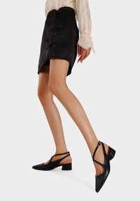 Scarpe slingback in pelle nera con punta affusolata, tacco basso a blocco e cinturini incrociati. Texture liscia, design minimalista.
