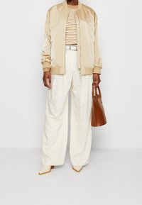 Chaqueta bomber de satén beige sobre una blusa de punto a rayas, combinada con pantalones de pernera ancha blancos a rayas y tacones beige puntiagudos, sosteniendo un bolso marrón.