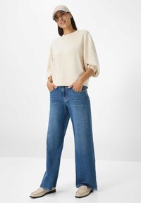 Licht beige cropped sweatshirt met oversized mouwen, gecombineerd met wijde blauwe jeans en lichtbruine loafers, en een bijpassende pet.