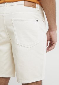 Pantalones cortos de mezclilla beige claro con un diseño clásico de cinco bolsillos, que presentan una etiqueta de cuero en la cinturilla y detalles de costura en contraste.