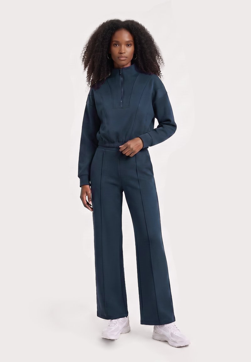 Femme aux cheveux bouclés portant un sweat-shirt zippé bleu marine et un pantalon assorti avec des baskets blanches, debout devant un fond uni.