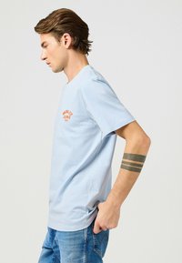 Ljusblå bomullströja med rund halsringning, korta ärmar och orange broderad text på bröstet. Slim fit-design med sidsprund.