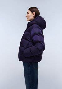 Fioletowa kurtka puchowa z kapturem, z pikowanym wzorem i czarną łatą z logo. W połączeniu z ciemnoniebieskimi dżinsami, ukazana w profilu bocznym.