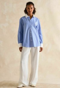 Blue striped button-up shirt with white cuffs and collar, paired with loose white linen trousers and light shoes. Simple embroidery detail on shirt.