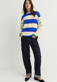Gestreepte trui in blauw, crème en groen; geribbelde halslijn en manchetten. Gecombineerd met losse spijkerbroek in donkerdenim en zwarte schoenen.