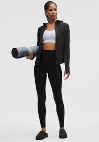 Fekete cipzáras dzseki egy fehér crop toppal, fekete magas derekú leggings és fekete slip-on cipők. Kényelmesen tart egy döntött dizájnú jógaszőnyeget.