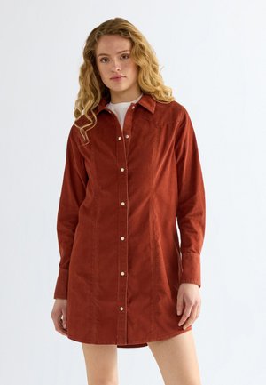 Donna con lunghi capelli biondi mossi che indossa un vestito camicia color ruggine sopra una maglia bianca, in piedi davanti a uno sfondo bianco uniforme.