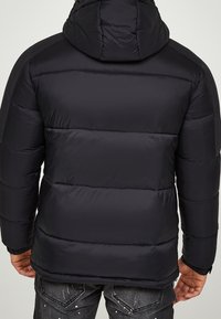 Mand iført en sort polstret hættetrøje og mørke jeans med malersplats detaljer, set fra baggrunden mod en ensfarvet baggrund.