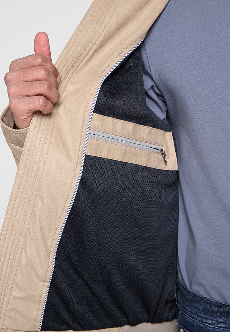 Veste beige avec un intérieur en maille noir et une poche zippée. La veste présente des coutures visibles et une texture de tissu léger.