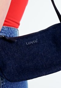 Mörkblå denim crossbody-väska med dragkedjestängning, har "Levi's"-logotypen i svart söm på ena sidan. Slät textur.