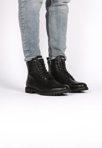 Botas de cuero negro hasta el tobillo con puntera redonda, cordones frontales, cremallera lateral y suela robusta. Combinadas con jeans de mezclilla azul claro.