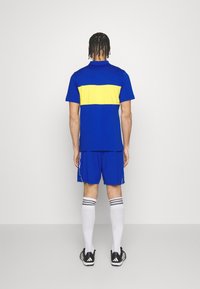 Modrý atletický outfit skládající se z trička s krátkým rukávem a žlutým pruhem, odpovídajících kraťasů, bílých ponožek s černými pruhy a černých bot.