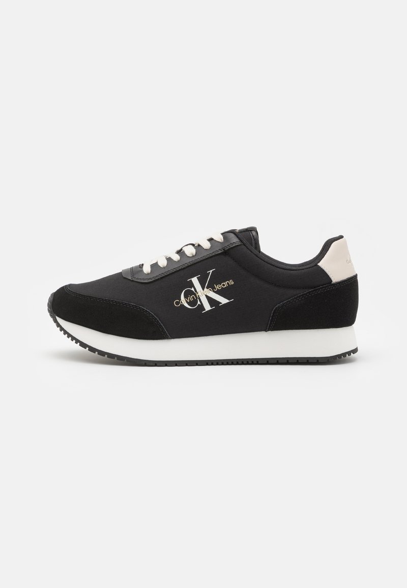 Zapatilla deportiva negra con superiores de tela y ante, suela blanca, detalles beis en el talón y un logotipo en blanco y dorado en el lateral.