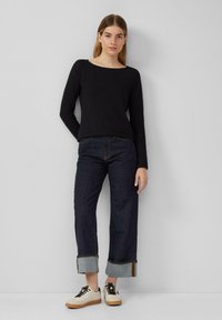 Black long-sleeve top, relaxed fit, paired with dark blue cropped jeans featuring turned-up hems, worn with cream sneakers and black laces.
