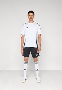 Bijela sportska majica s crnim detaljima, u kombinaciji s tamnim hlačicama, bijelim čarapama s tri crne pruge i bijelim kopačkama za nogomet.