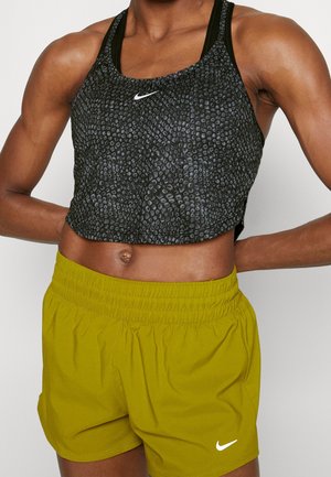 Athletische Person trägt ein schwarzes, strukturiertes Nike Crop-Top und gelbe Nike-Shorts, die Hände ruhen im unteren Rücken.