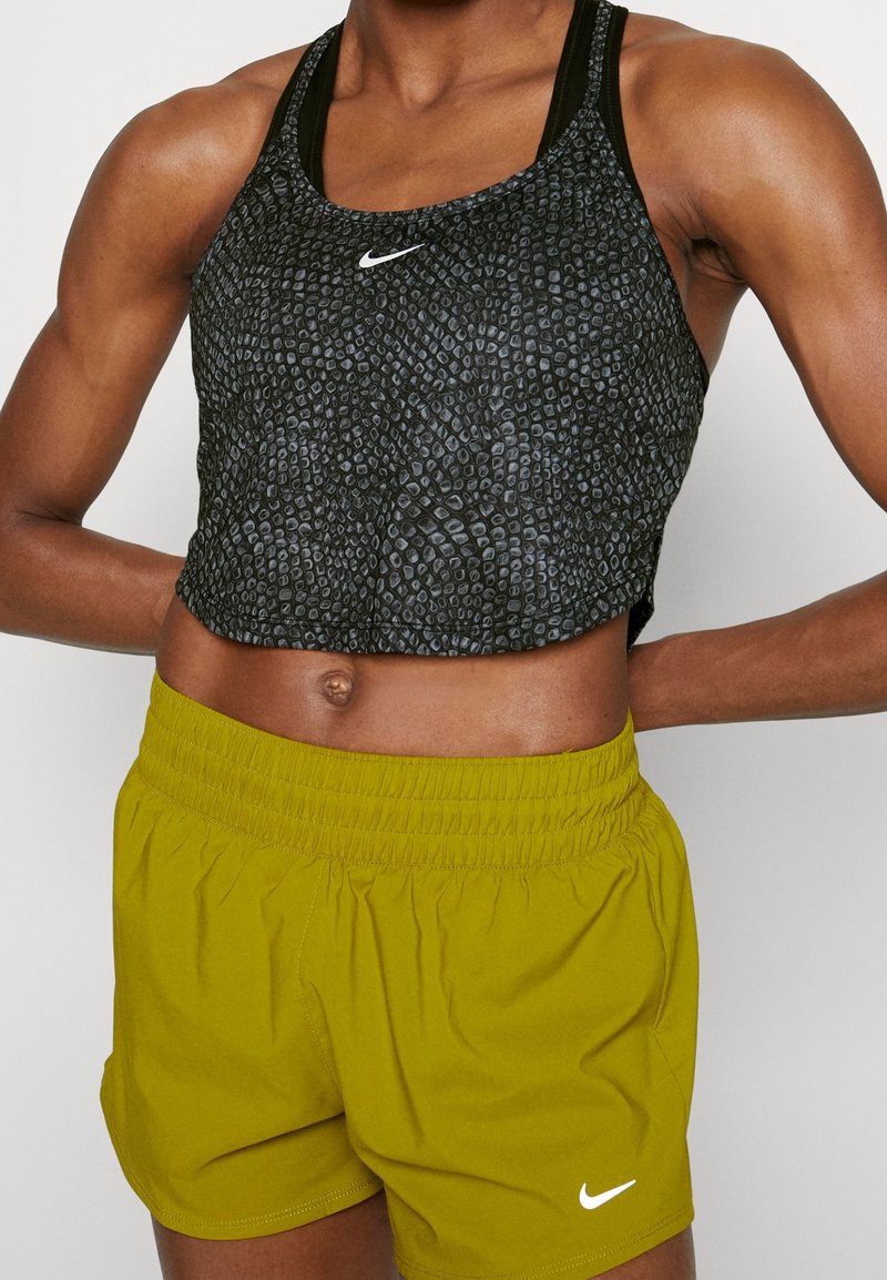 Personne athlétique portant un crop top noir texturé Nike et un short jaune Nike, mains reposant sur le bas du dos.