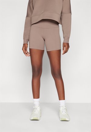 Modelka nóg nosząca beżowe, sportowe szorty z wysokim stanem oraz pasującą bluzę z długim rękawem, do tego białe skarpetki i trampki, stojąca wewnątrz pomieszczenia.
