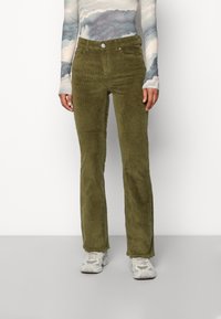 Persoon in olijfgroene corduroy broek, een lange mouwen patroon top en witte sportschoenen die tegen een eenvoudige achtergrond staat.