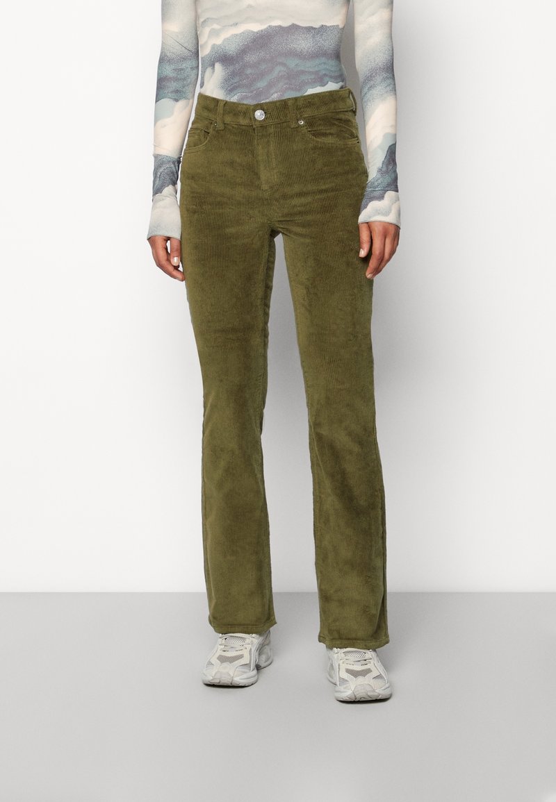 Persoon in olijfgroene corduroy broek, een lange mouwen patroon top en witte sportschoenen die tegen een eenvoudige achtergrond staat.