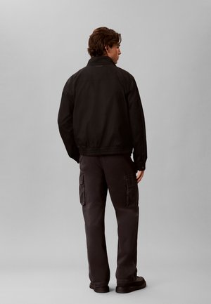 Mann steht mit dem Rücken zur Kamera, trägt eine schwarze Jacke und dunkle Cargohosen mit Seitentaschen vor einem einfarbigen grauen Hintergrund.