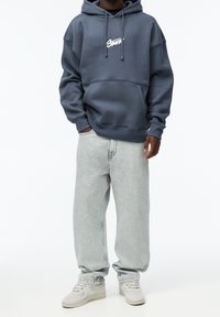 Tmavě modrá mikina s kapucí a přední kapsou, doplněná o světle šedé volné džíny a béžové tenisky. Hladký povrch látky.