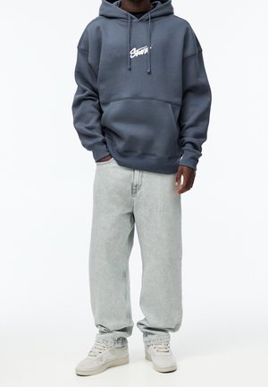 Tmavě modrá mikina s kapucí a přední kapsou, doplněná o světle šedé volné džíny a béžové tenisky. Hladký povrch látky.