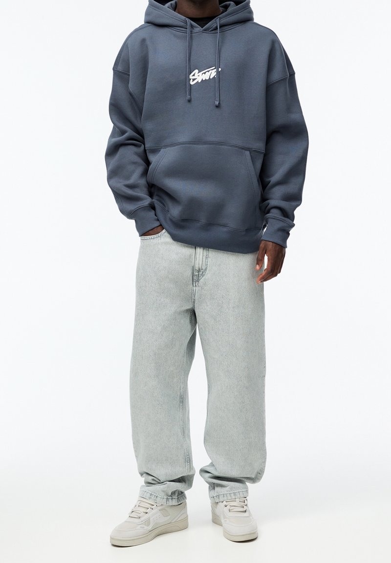 Tmavě modrá mikina s kapucí a přední kapsou, doplněná o světle šedé volné džíny a béžové tenisky. Hladký povrch látky.
