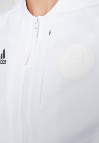 Gros plan d'une veste blanche zippée avec le logo Adidas sur la poitrine gauche et un emblème circulaire embossé sur la poitrine droite.