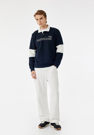 Felpa blu navale con dettagli crema e testo "FEARLESS IN ACTION", abbinata a pantaloni bianchi e sneaker nere. Design con colletto incrociato.